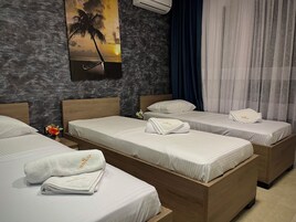 Habitación de hotel con tres camas, cada una con ropa blanca y toallas, aire acondicionado de pared, un cuadro de un atardecer sobre el océano y una pared oscura con textura.