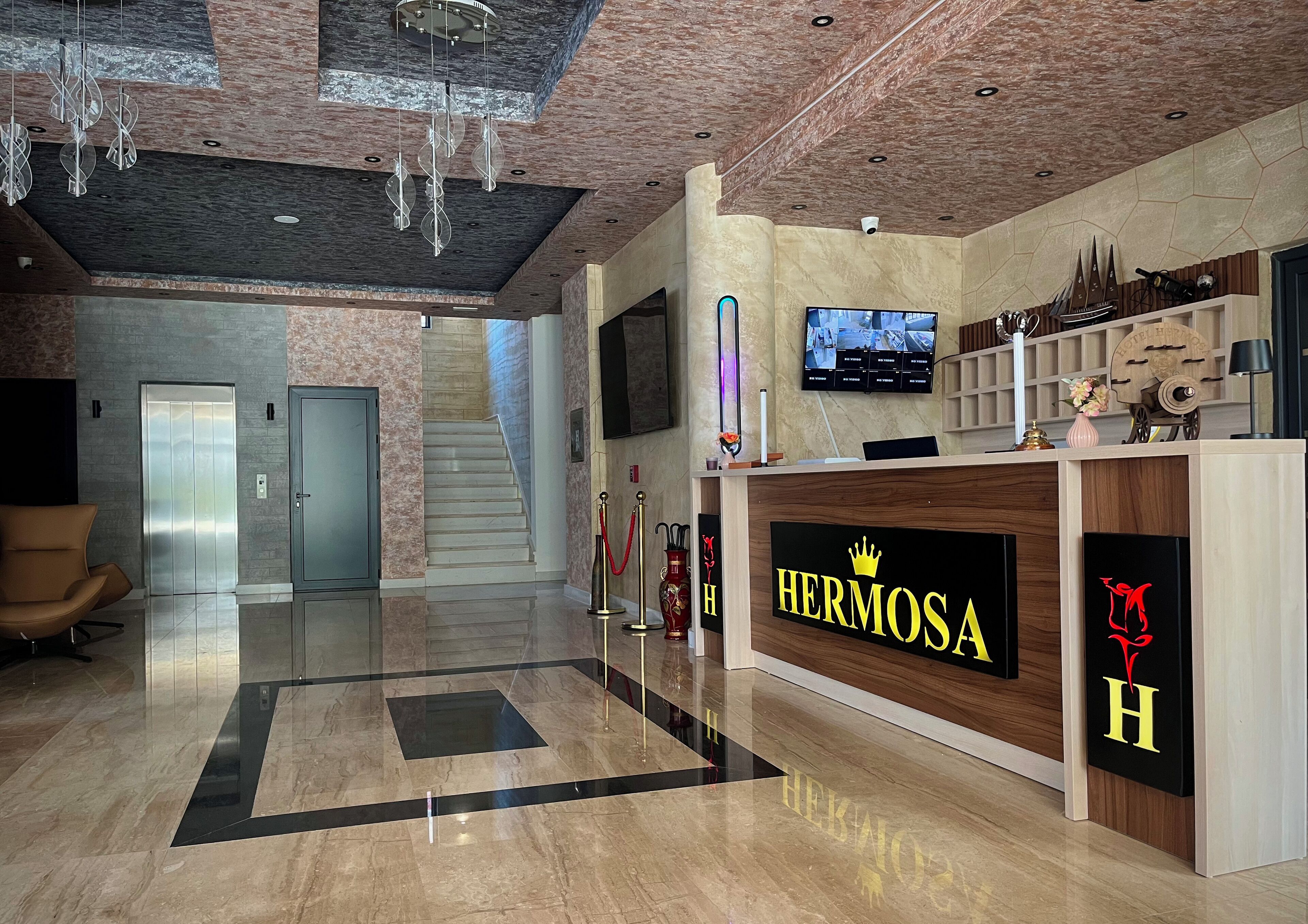 A hotel lobby with a reception desk labeled "HERMOSA," a staircase, and a modern chandelier.