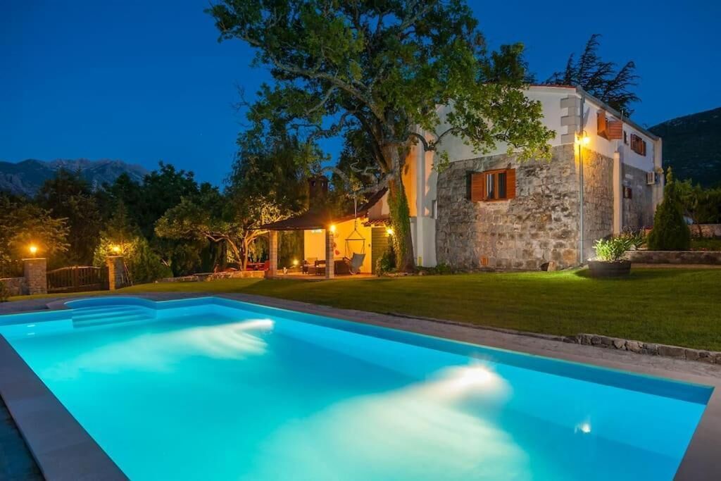 A poolside view of a stone building with a swimming pool, surrounded by trees and a mountainous backdrop.