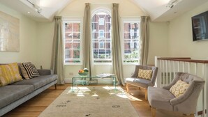 A living room with a sofa, armchair, glass coffee table, and a view of a brick building through large windows. - Charming Notting Hill Mews House (London)