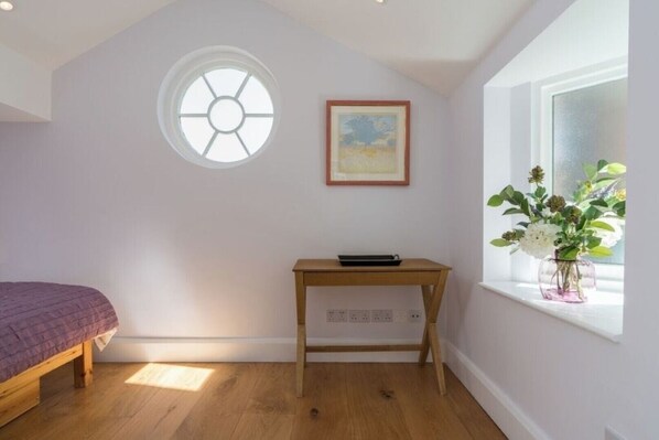A bedroom with a bed, a small wooden table, a window with a view of flowers, and a ceiling light.