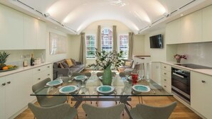 A modern kitchen with a glass dining table, white cabinets, and a large window with curtains. - Charming Notting Hill Mews House (London)
