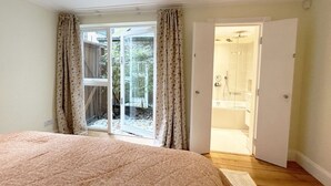 A bedroom with a large bed, a sliding glass door leading to a balcony, and a bathroom visible through an open door. - Charming Notting Hill Mews House (London)