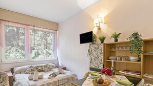 A cozy room with a bed, a dining table with a floral tablecloth, a television, and a bookshelf with plants. - Sonia Sweet Love With Shared Pool (Gardone Riviera)