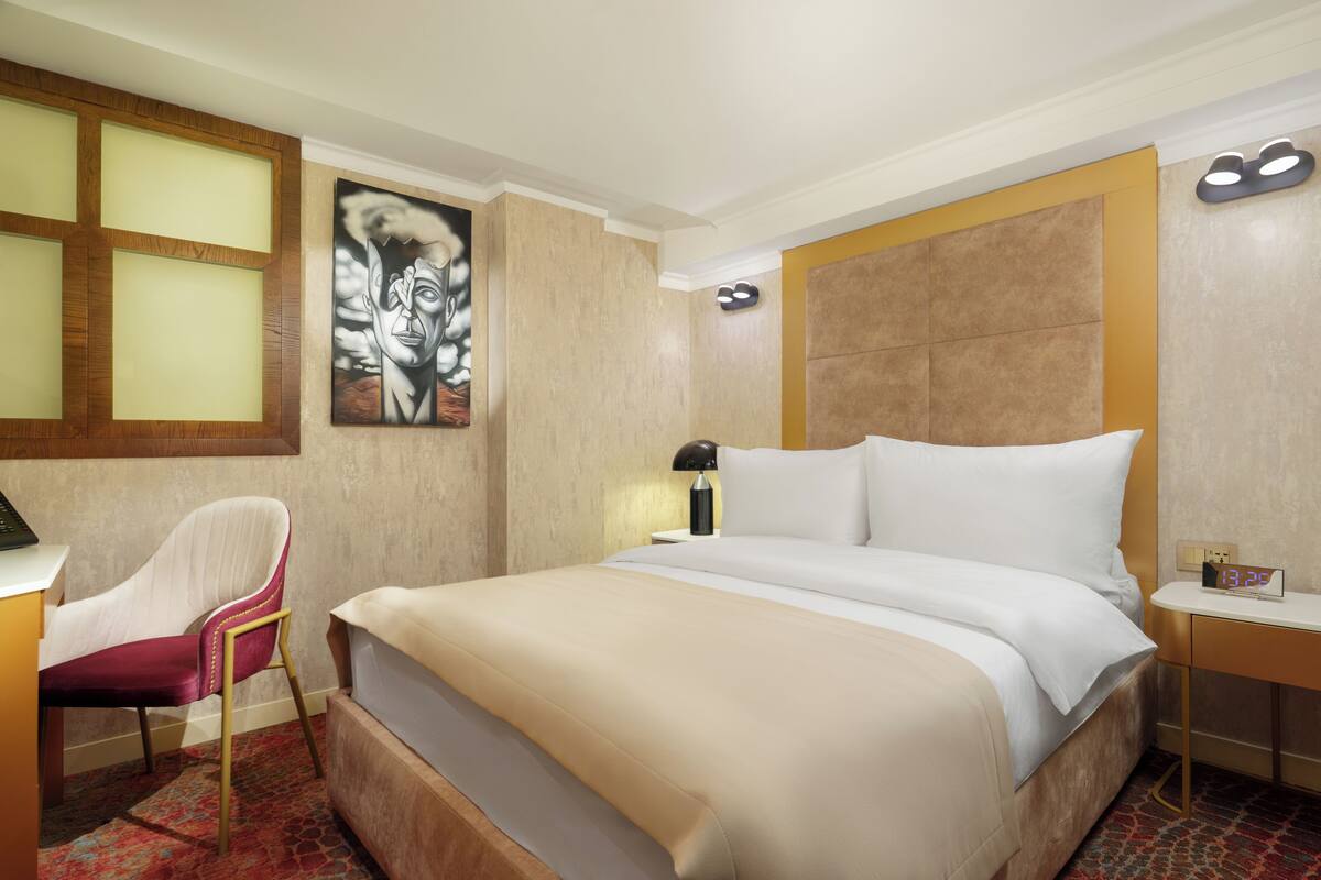 A hotel room with a bed, a desk, a chair, a painting on the wall, and a nightstand with a lamp.
