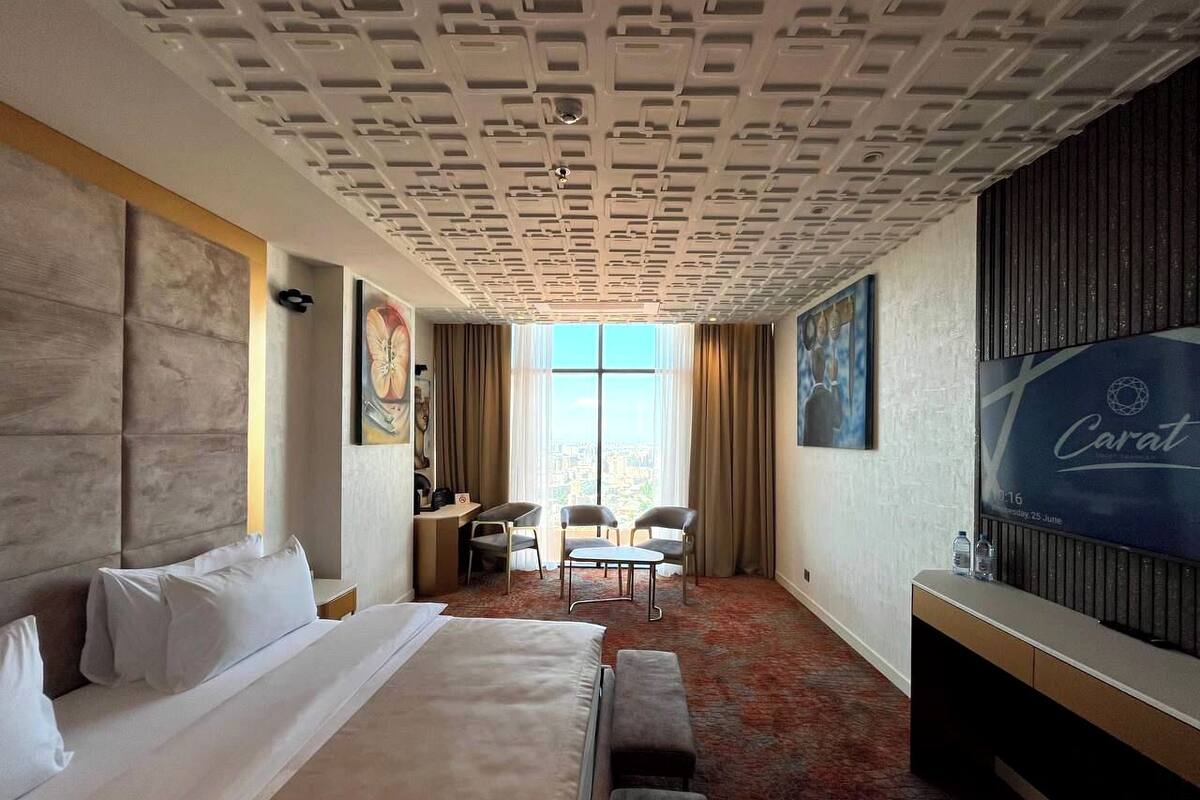 A hotel room with a large bed, a desk with two chairs, a TV, and a large window with curtains.