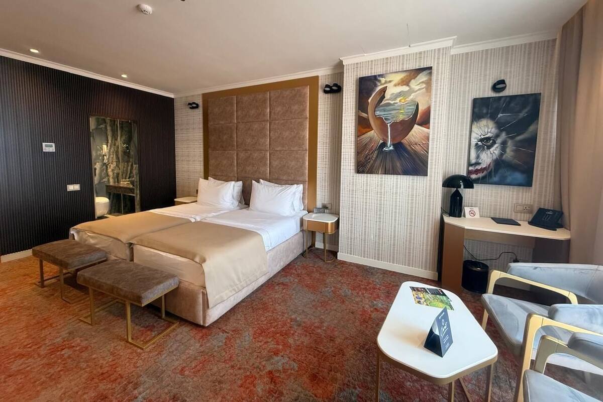 A hotel room with a bed, bedside tables, a desk, a chair, a bench, and artwork on the walls.