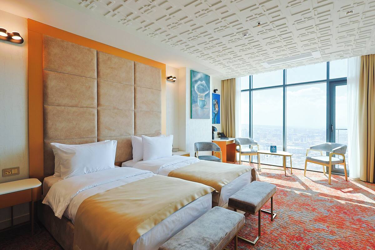 A hotel room with two beds, a desk, and a large window offering a city view.