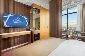 Une chambre d’hôtel dotée d’un grand téléviseur affichant le logo Carat, d’un lit, d’un bureau avec une machine à café et offrant une vue sur le paysage urbain grâce à de grandes fenêtres.