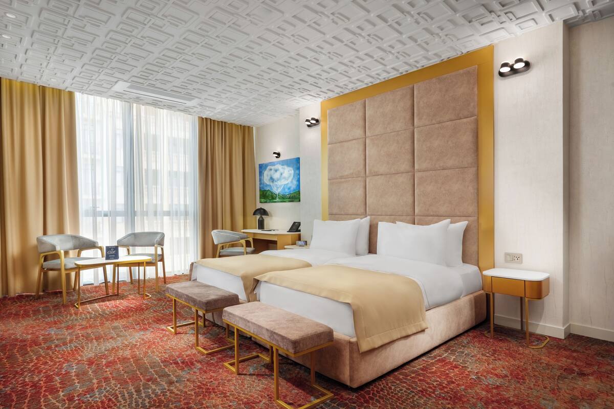 A hotel room with a large bed, two bedside tables, a desk with a chair, a bench, and a large window with curtains.
