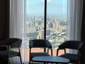 A room with a large window offering a cityscape view and a mountain in the distance. There are two chairs and a small table in the foreground.