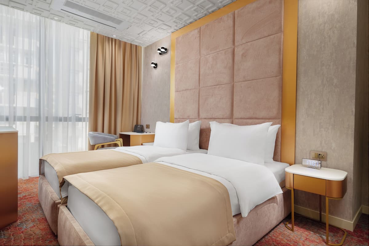 A hotel room with two beds, a desk, a chair, a nightstand, and a large window with curtains.