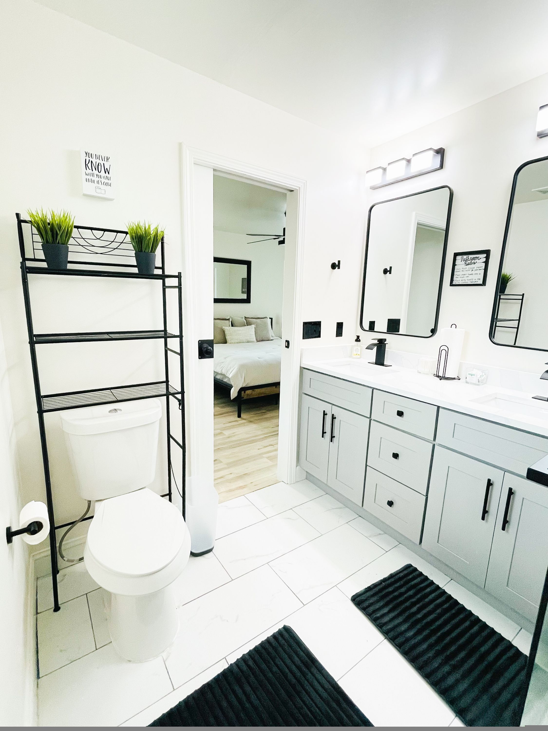 A modern bathroom with a toilet, sink, and mirror. There is a black metal shelf with plants and a black rug on the floor.