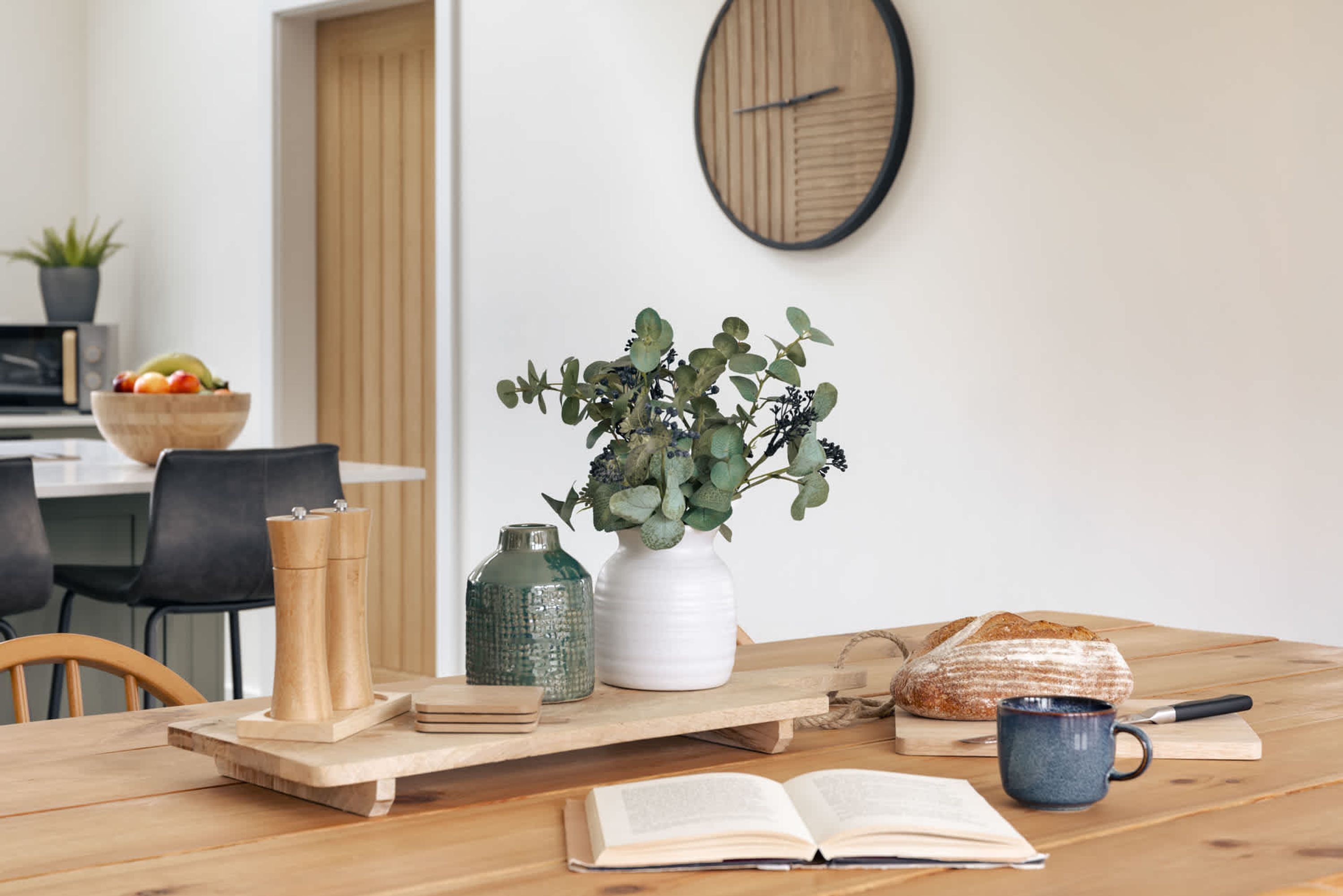 Una mesa de madera con un libro, una taza, un pan y un florero con plantas verdes.