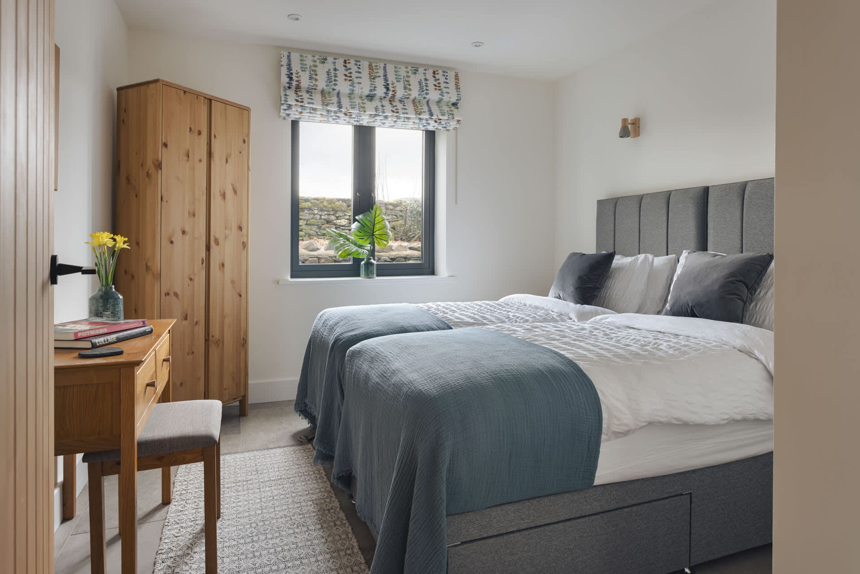 A bedroom with two beds, a desk with a chair, a wardrobe, and a window with a view.