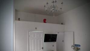 A room with a white door, a black folding chair, a television mounted on the wall, a small cabinet with a fan, and a chandelier. - Apartmentsleeps4-west Kensington-petsok-parking (London)