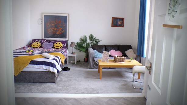 A cozy bedroom with a bed, a sofa, a small table, and a chair. There is a window with curtains, a wall with a picture, and a door with a handle. - Apartmentsleeps4-west Kensington-petsok-parking (London)