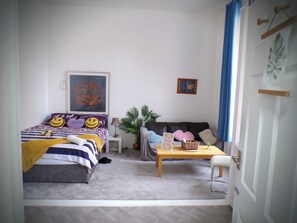 Ein gemütliches Schlafzimmer mit Bett, Sofa, kleinem Tisch und Stuhl. Ein Fenster mit Vorhängen, eine Wand mit einem Bild und eine Tür mit Griff.
