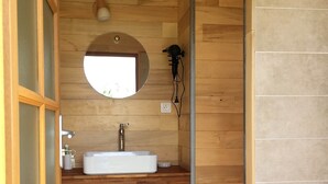 Une salle de bain avec un plancher en bois, un miroir rond, un lavabo blanc et un luminaire.