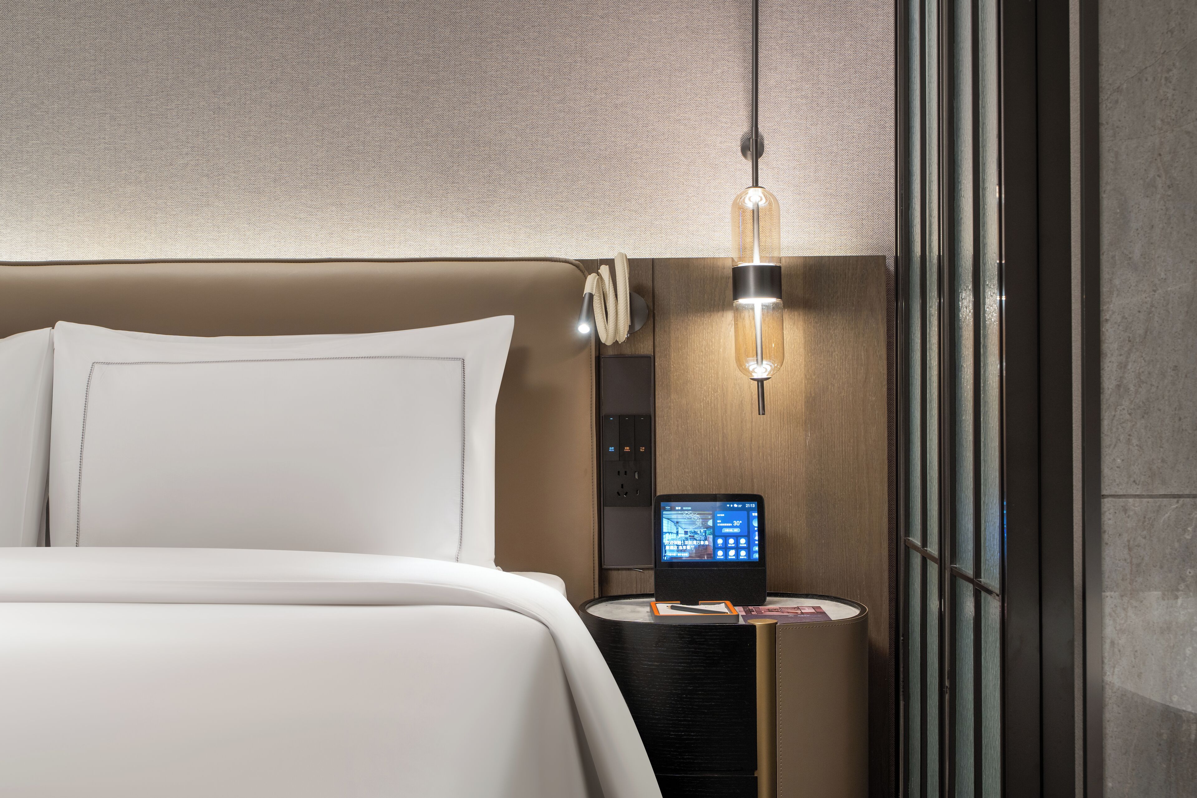 Habitació d'hotel moderna amb un llit gran, una taula de nit amb un panell de control digital, una làmpada i vista exterior.