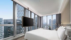 Une chambre d’hôtel moderne dotée d’un grand lit, d’un téléviseur à écran plat, d’une petite table avec une lampe et offrant une vue sur la ville grâce à de grandes fenêtres.