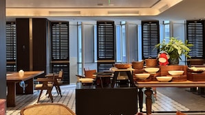 Ein moderner Essbereich mit mehreren Tischen und Stühlen, großen Fenstern mit Jalousien und einem zentralen Tisch mit dekorativen Schalen und einer Topfpflanze.
