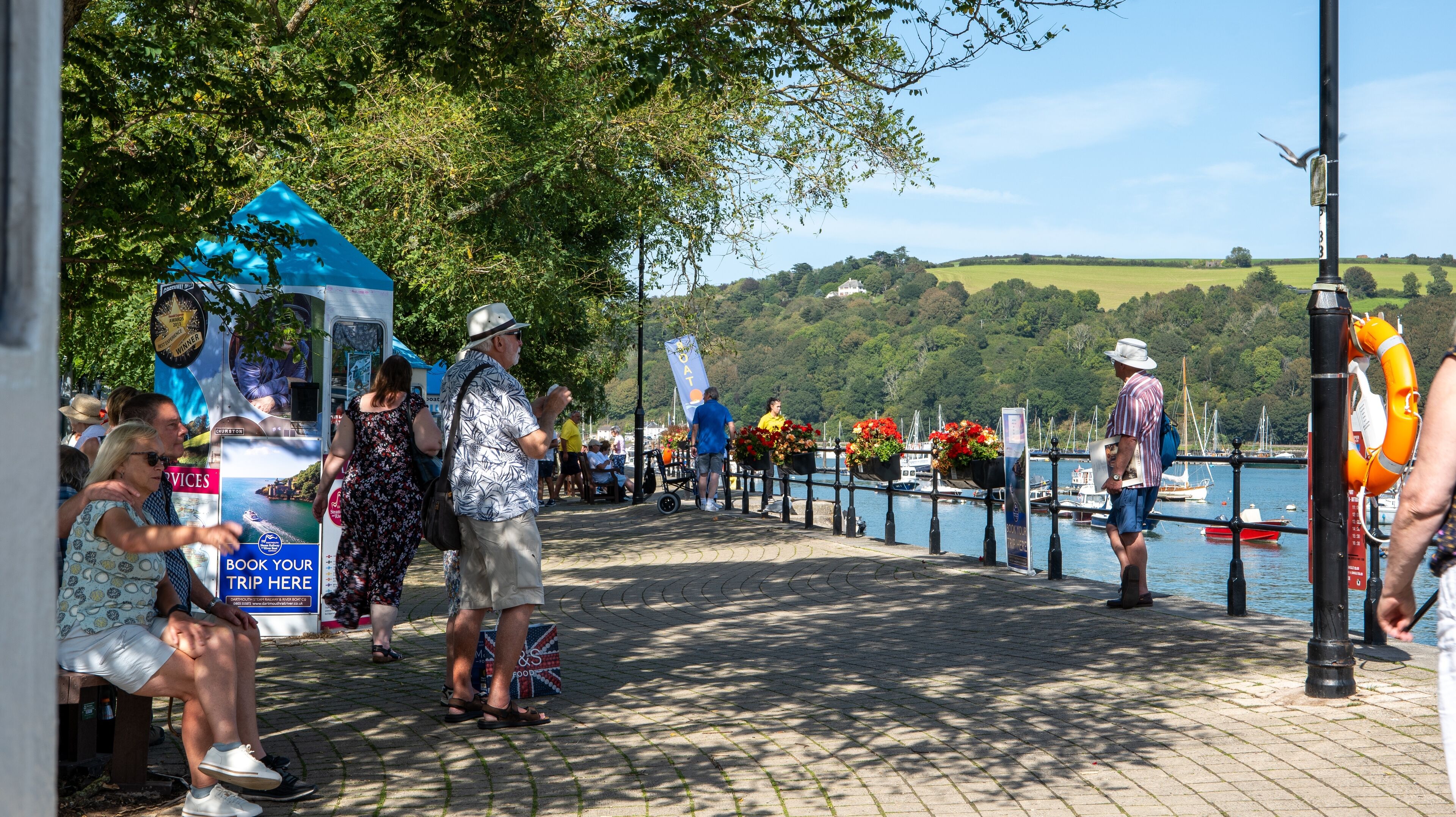 A waterfront promenade with a blue tent, people walking, and boats in the background.