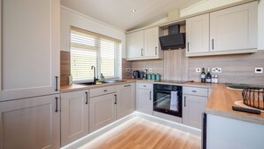 A modern kitchen with light wood cabinetry, a built-in oven, and a window with blinds. - Countryside Lodge Retreat on Golf Course (Blackawton)