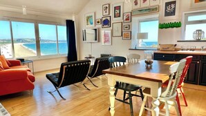 A spacious living room with a large window overlooking the beach, a dining area with a wooden table and chairs, and a kitchen with modern appliances and artwork on the walls. - ​Beachfront Cottage with Sea Views & Coastal Walks​ (Torcross)