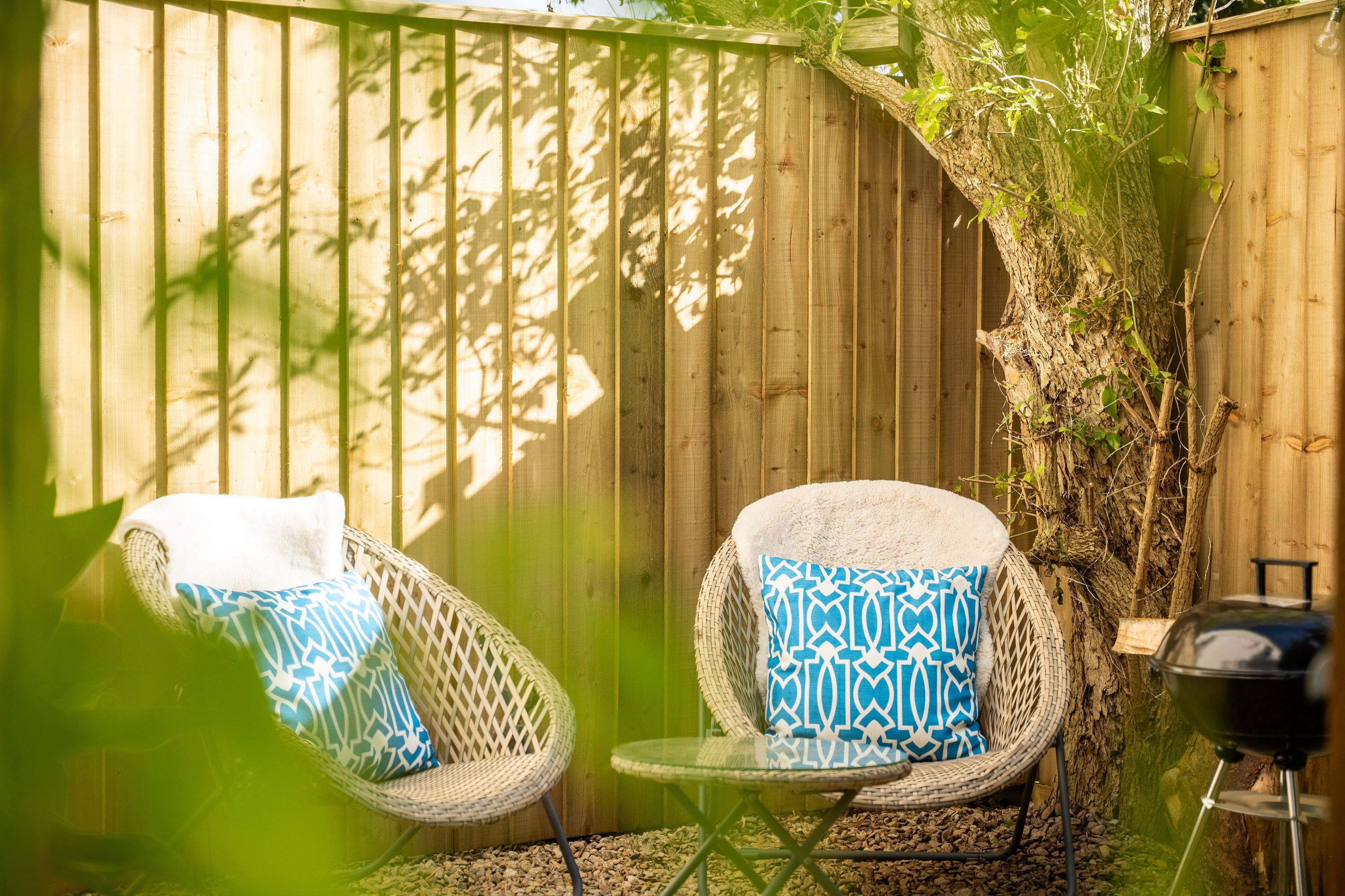 Two wicker chairs with patterned cushions, a small glass table, and a barbecue grill, all set against a wooden fence with climbing plants.