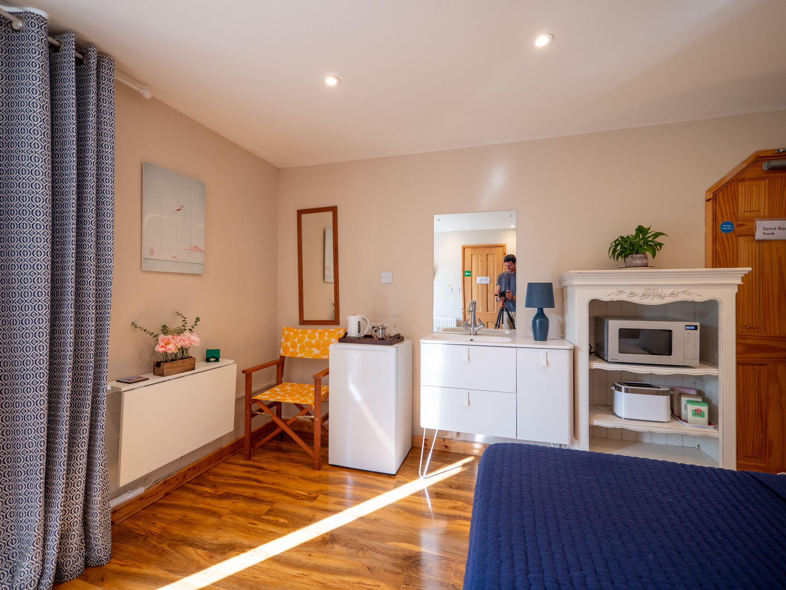 A hotel room with a wooden floor, a bed, a desk with a chair, a mirror, a microwave, and a plant.