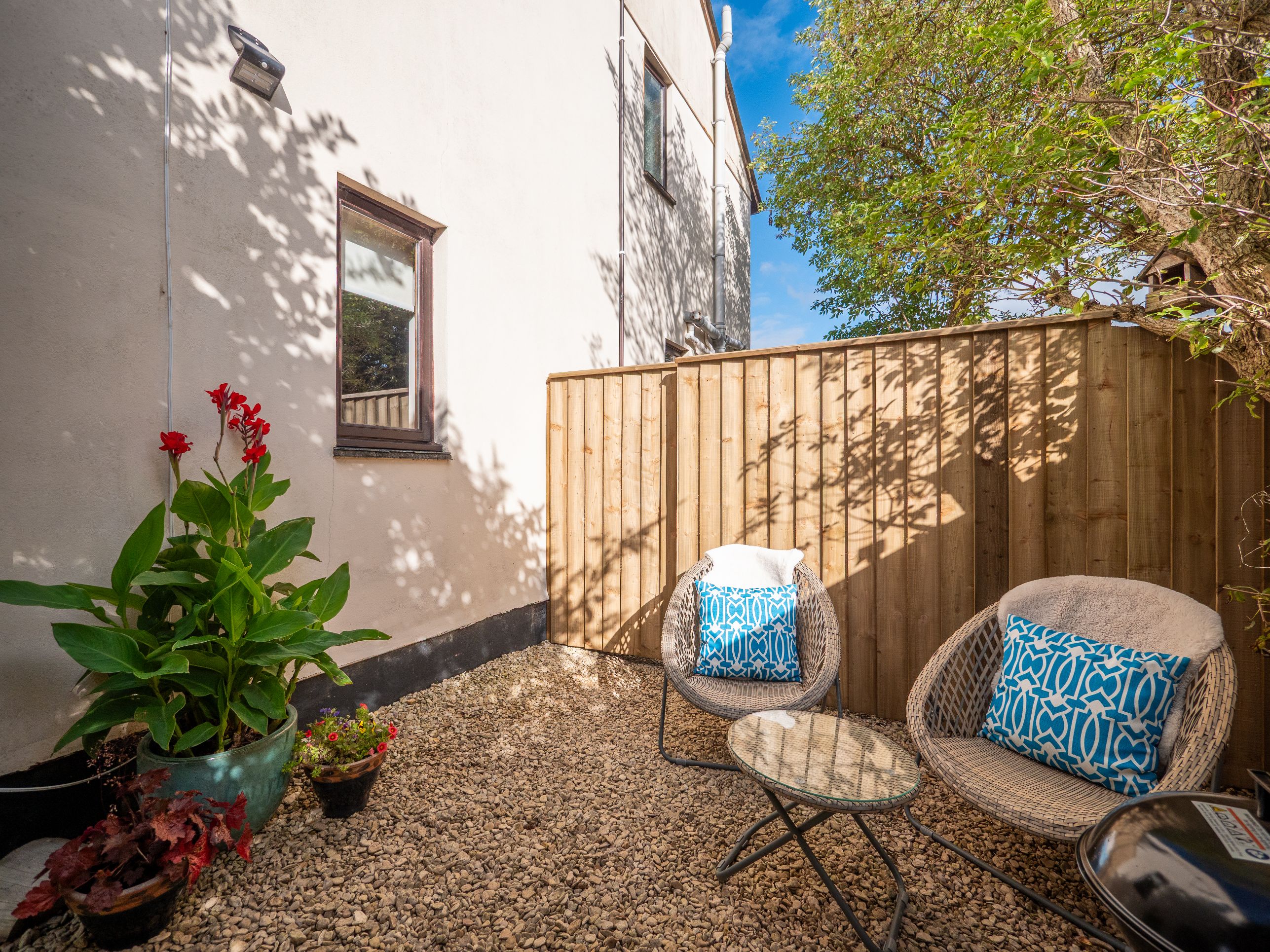 A patio with a wooden fence, a potted plant, and two wicker chairs with cushions.