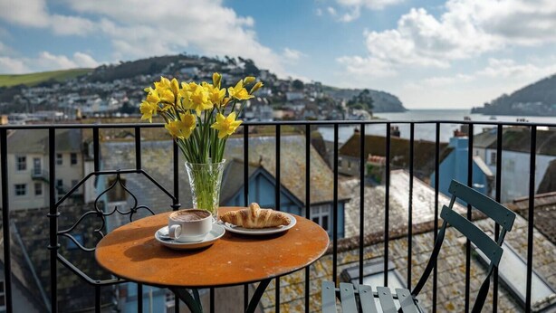 A balcony with a table set for a meal, a vase of yellow flowers, and a view of a coastal town.