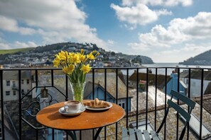 A balcony with a table set for a meal, a vase of yellow flowers, and a view of a coastal town. - River View Apartment with Parking & Balcony (Dartmouth)