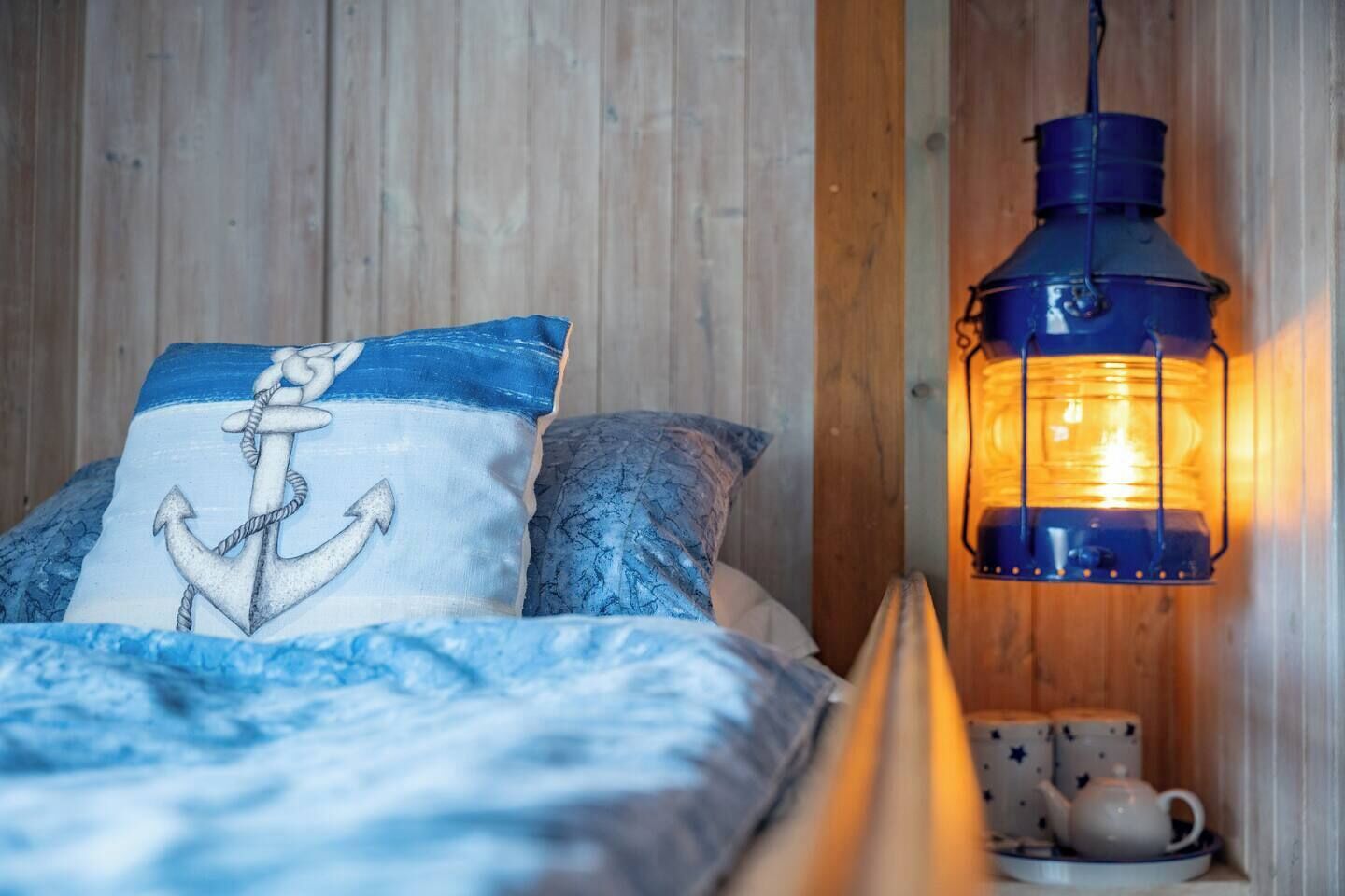 A bed with a blue and white nautical-themed pillow, a patterned pillow, and a blue blanket. A blue lantern-style light fixture is mounted on the wall beside the bed.