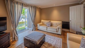 Un salon avec un canapé beige, un meuble en bois foncé, une télévision et une grande fenêtre avec des rideaux.