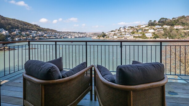 A balcony with wicker chairs and cushions, overlooking a coastal town and a body of water. - Stylish Waterfront Home With River Dart Views (Dartmouth)