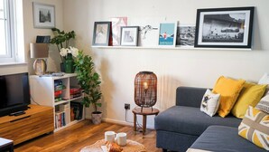 A living room with a grey sofa, a TV on a wooden stand, a wicker ottoman with croissants, a potted plant, and a wall with framed pictures.