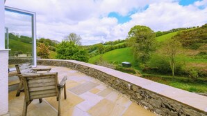 A stone patio with a wooden bench, overlooking a green valley and hills.