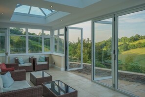 A spacious sunroom with large glass doors and windows, wicker furniture, and a view of a green landscape. - 18th-Century Farmhouse with Countryside Views (Blackawton)