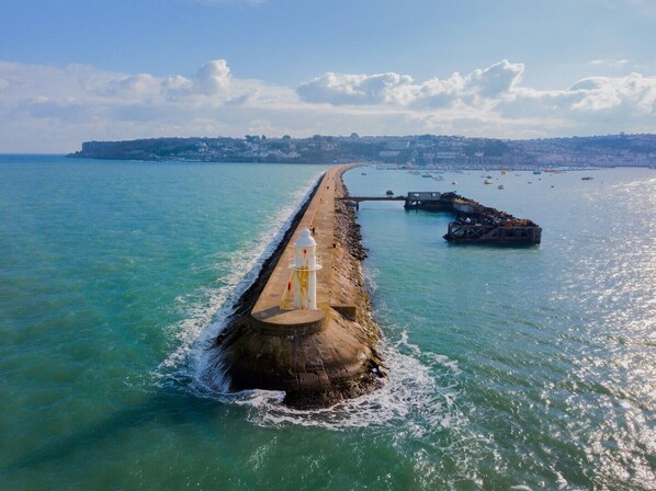 Un muelle costero se extiende hacia el mar, con un faro al final, rodeado de aguas tranquilas y una costa distante.