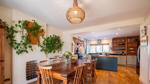 A wooden dining table with chairs, a hanging light fixture, and a kitchen area with cabinets and a refrigerator. - Farmhouse with Garden & Countryside Views (Totnes)
