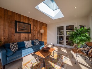 A modern living room with a wooden wall, a blue sofa, a wooden coffee table, a patterned rug, and a large window. - Farmhouse with Garden & Countryside Views (Totnes)