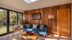 A modern living room with a blue sofa, wooden panel walls, a large window, and a skylight. - Farmhouse with Garden & Countryside Views (Totnes)