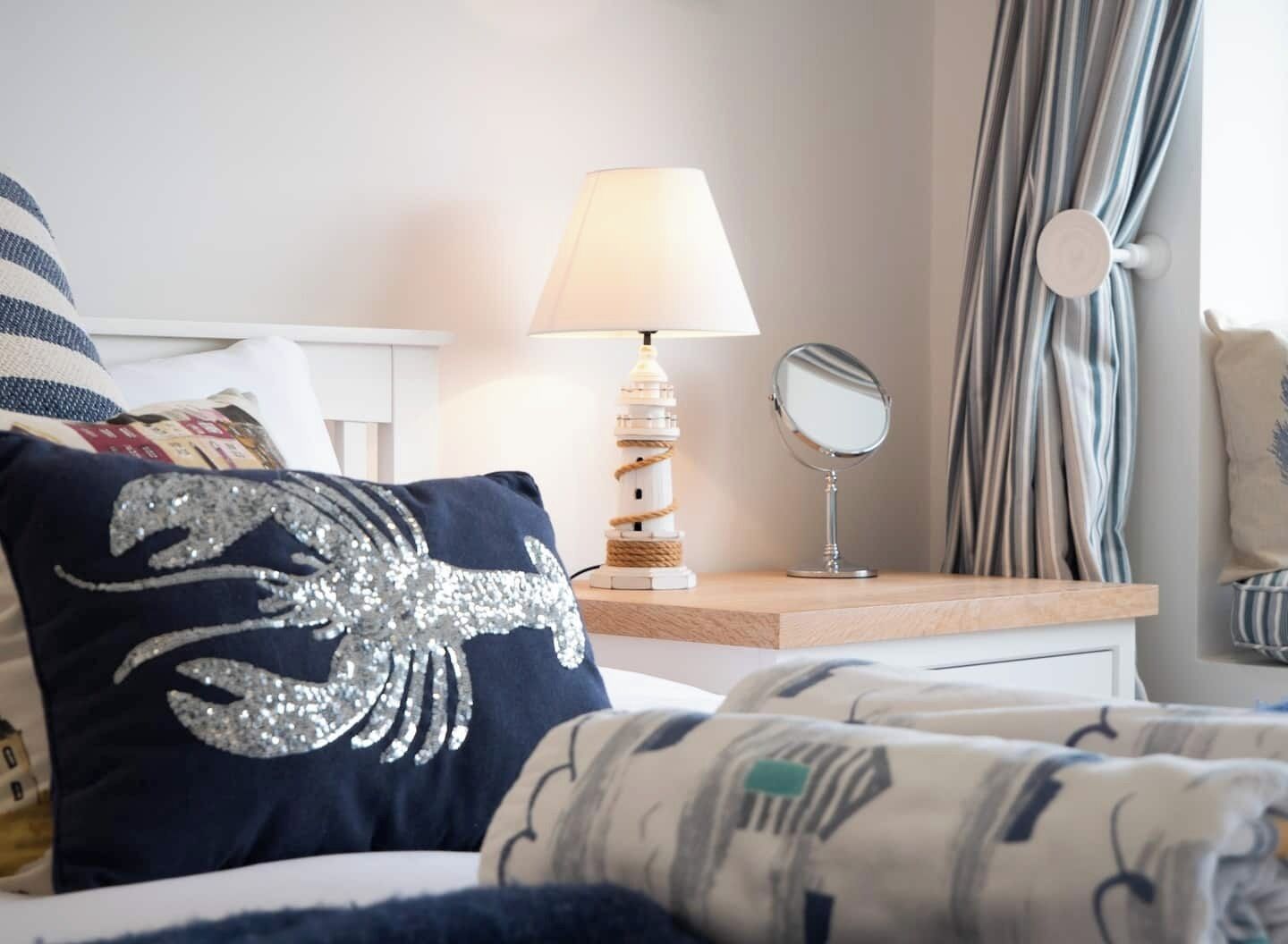 Un lit bien fait, agrémenté d’un coussin décoratif, d’une lampe de chevet, d’un miroir rond et d’un rideau à rayures.