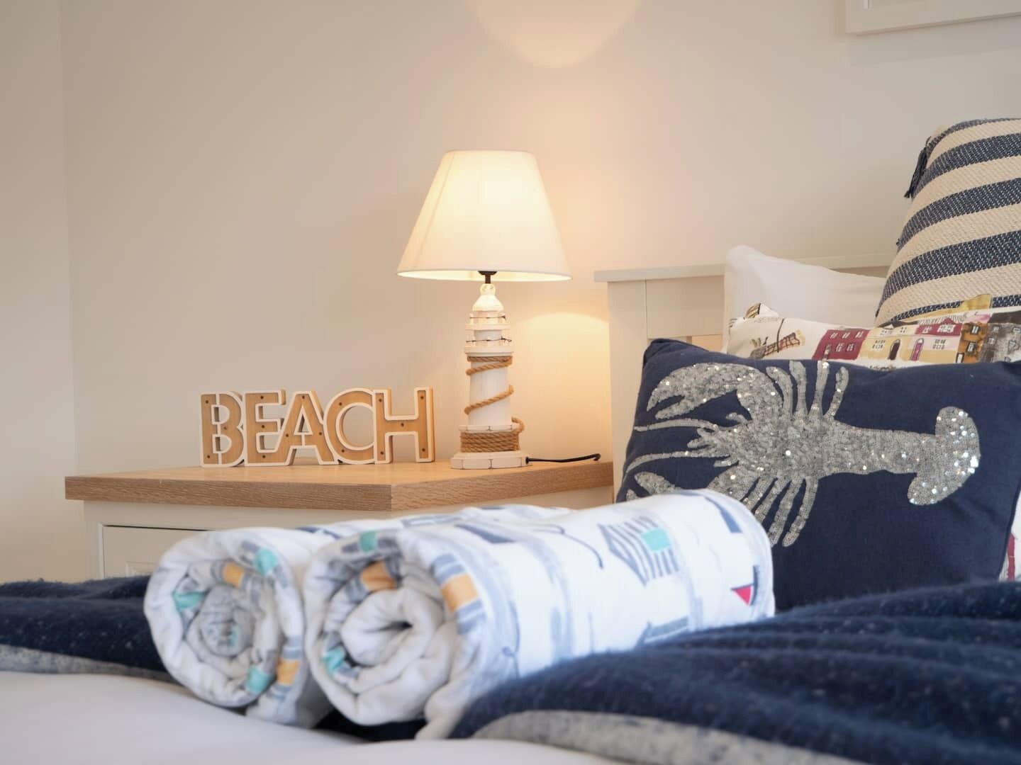 A neatly made bed with a rolled-up blanket, a lamp, a 'BEACH' sign, and a decorative pillow with a seahorse design.