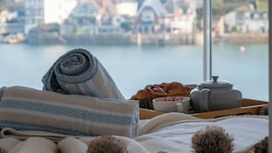 Ein Bett mit gerolltem Handtuch, ein Tablett mit Brot, ein Teekessel und eine Tasse mit Blick auf einen malerischen Hafen und Häuser.