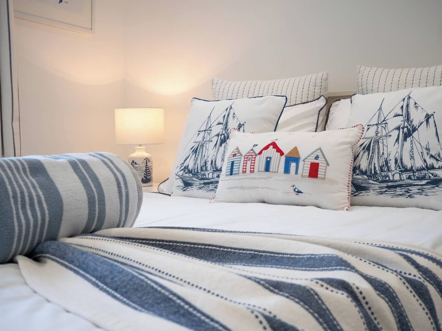 Una cama bien hecha con ropa de cama a rayas, una lámpara y almohadones decorativos con motivos marinos.