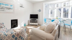 A living room with a beige sofa, patterned armchairs, a TV on a white stand, a fireplace, and a dining area with blue chairs. - Charming Apartment with Garden & Parking (Dartmouth)
