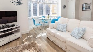 A modern living room with a white sofa, blue chairs, a flat-screen TV, and a dining area with a table and chairs. - Charming Apartment with Garden & Parking (Dartmouth)